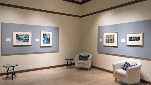 View of gallery with prints on the wall, and two armchairs and side tables