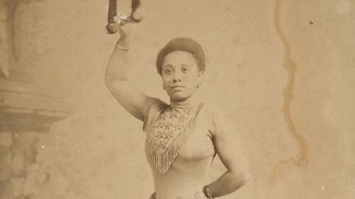 Sepia photo of a petite Black woman wearing a decorated leotard, standing and holding onto a trapeze with her right arm