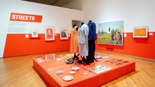 Gallery view with paintings and 3 Soviet-era outfits and other objects on a red platform; wall text says “Streets”