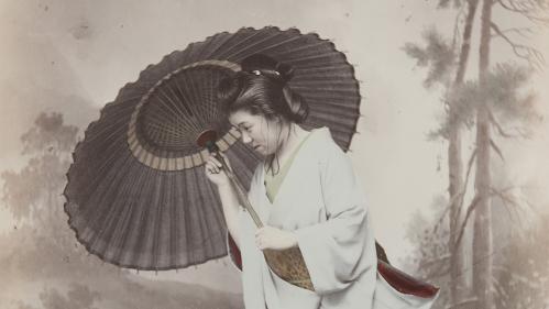 Handcolored studio photo of young Japanese woman holding a parasol against a gust of wind