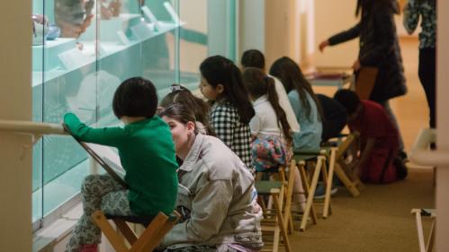 Children sit before a display case drawing on clipboards