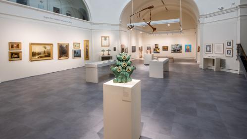 Large gallery room with white walls and gray floor tiles. Paintings hang on the walls and in the foreground is a green ceramic sculpture on a pedestal.