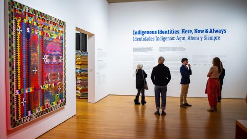 people standing at entrance to exhibition, under the title, Indigenous Identities: Here, Now & Always, on the wall