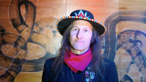 Neal Ambrose-Smith, a Native American man, stands in front of a patterned wall while wearing a decorated hat, his long hair falls over his shoulders. He has on a red handkerchief and a black jacket with a variety of pins on the lapel.