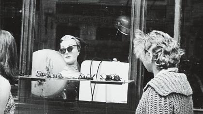 black and white photo of two women looking in the window of an eyeglass shop
