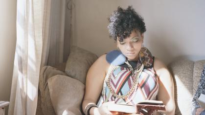 Photo of a young Black woman wearing a patterned dress, seated on a couch besides a window and reading
