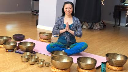 Photo of young Asian woman seated with palms pressed together, surrounded by Tibetan singing bowls of various sizes