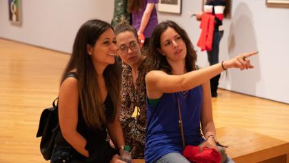 3 young women sit on a bench in a gallery looking at art out of the camera's view; one points