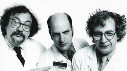 three white middle aged men in lab coats stare at the camera