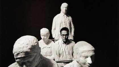 black and white photo of a white man with glasses sitting among plaster-cast figures