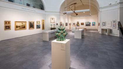 Large gallery room with white walls and gray floor tiles. Paintings hang on the walls and in the foreground is a green ceramic sculpture on a pedestal.