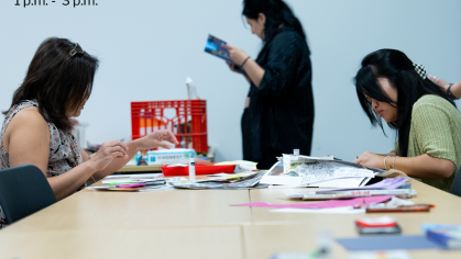 Two people sit at a table making art while a third person stands reading a packet.