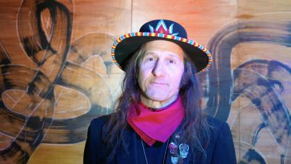 Neal Ambrose-Smith, a Native American man, stands in front of a patterned wall while wearing a decorated hat, his long hair falls over his shoulders. He has on a red handkerchief and a black jacket with a variety of pins on the lapel.