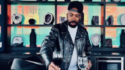 A silhouetted bar with liquor bottles and a green hue on the back wall. Behind the bar is Keith Jones II wearing a black suit jacket, white shirt, and a black brimmed hat turned backward. He is offering a wine glass full of red wine to the camera.