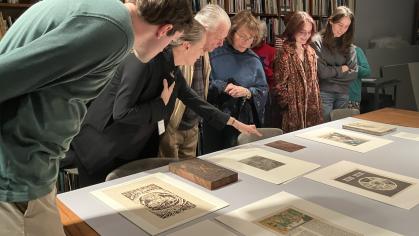 A large wood table had several matted prints laying on it. The prints are etchings and have been made in black ink. Standing over the table is a group of six people viewing the artworks. One of the points to a particular artwork while others look.