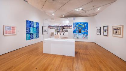 Museum gallery with wood floors and white walls. On the walls hang a variety of paintings that feature imagery of an elk crying, a parodied American flag, and protest text. In the center of the gallery is a large glass vitrine with a sculpture in the shape of an Indigenous war shirt. The shirt fabric has imagery of landscapes, people, and vehicles on it.