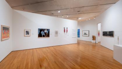 Gallery space with wood floors and white walls. On the walls are artworks varying in mediums: paintings, beadwork, sculpture.