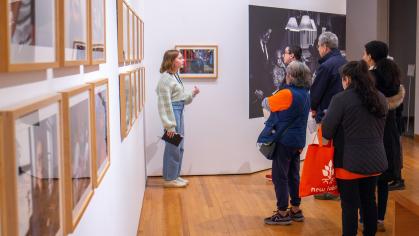 Gallery with white walls and wood floor featuring photographs framed along the wall behind glass. A tour guide speaks about the images to a groups of people dressed in winter clothing.