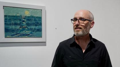 Wes Sherman wears a black collar shirt and stylish glasses. He has a bear and a shaved head, his hands hold a folded pamphlet. Sherman stands in front of a white wall that features a framed landscape painting.