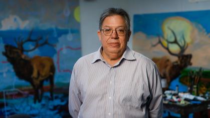 Artist Norman Akers stands in front of two paintings wearing a light blue collar shirt, glasses, and his hair tied back. His arms are behind his back. The two paintings in the background features elks amidst seas of garbage and devastated landscapes. The elks are crying out in what appears to be anguish.