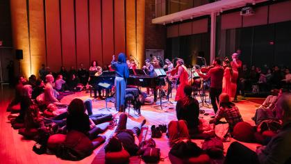 Concert hall lit in orange and pink light. Audience members seated on the floor surround a string group.