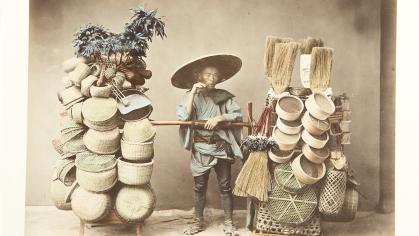 Handcolored studio photo of a young male Japanese vendor between two tall displays of baskets and brooms