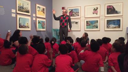 Children sit on the floor in an art gallery, listening to a storyteller standing in front of them