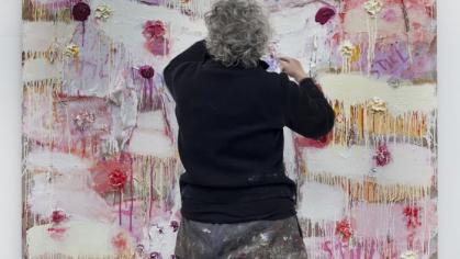Photo of Joan Snyder, an older woman dressed in jeans and black shirt, seen from behind painting an abstract canvas