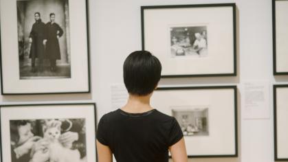 A woman with short dark hair with a black shirt & red skirt seen from behind, looking at photos in an exhibition