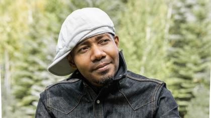 DJ Spooky smiling at the camera in front of him on a sunny day outside. He is seated and the background is out of focus green foliage.