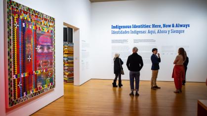 people standing at entrance to exhibition, under the title, Indigenous Identities: Here, Now & Always, on the wall