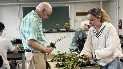 Two people, one elderly and the other in their mid to late twenties, grab craft materials from a table in a classroom.