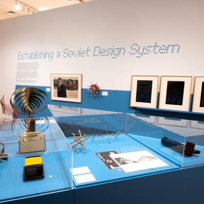 Gallery view of sculptures and household objects on a blue pedestal; wall text says “Establishing a Soviet Design System”