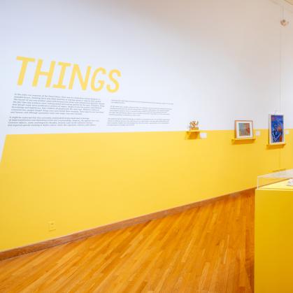 View of gallery with paintings and many small objects gathered on a large yellow pedestal; wall text says “Things”