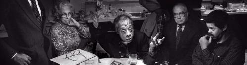 James Baldwin seated and leaning on left elbow at a table surrounded by people listening to him 