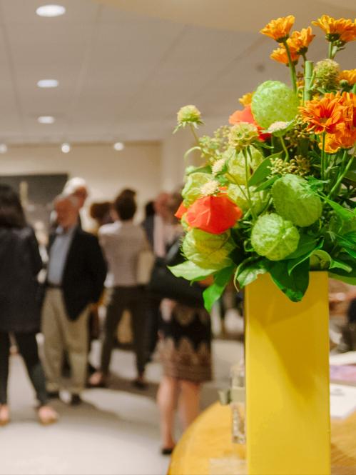 Opening reception guests in lobby with bright yellow vase of flowers at far right