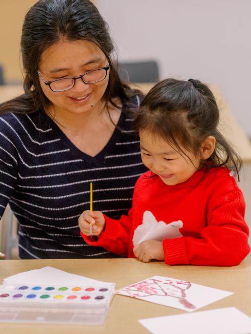 A child sits on a woman's lap. Both are smiling and painting with watercolors.