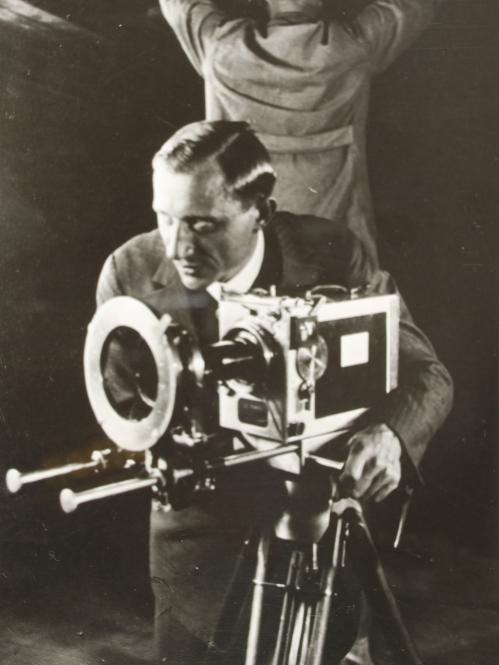 Black and white photograph of a cameraman crouched behind a large camera on a tripod 