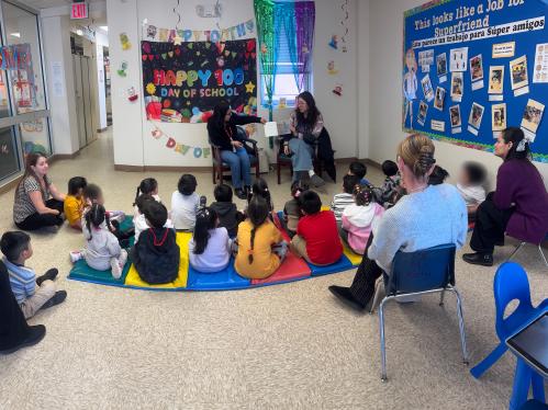 A group of children sit on mats on the floor while two hosts read from a picture book.