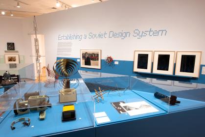 Gallery view of sculptures and household objects on a blue pedestal; wall text says “Establishing a Soviet Design System”