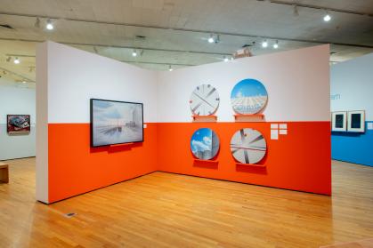 Gallery view of 5 paintings (1 rectangular, four round) of cityscapes on wall with red band at bottom