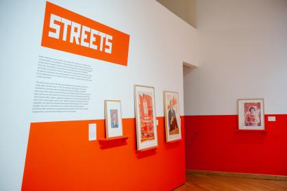 Gallery view with 4 prints on wall painted red on bottom, white on top; wall text says “Streets”
