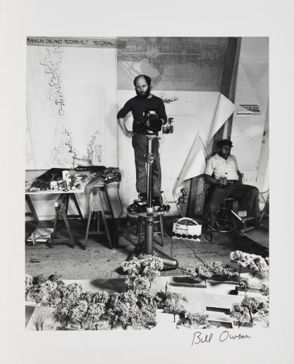 Black & white photo of man standing on a stool to photograph an architectural model. Another man sits on a chair to the side.