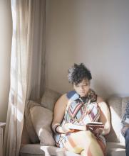 Photo of a young Black woman wearing a patterned dress, seated on a couch besides a window and reading