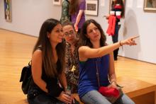 3 young women sit on a bench in a gallery looking at art out of the camera's view; one points