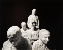 black and white photo of a white man with glasses sitting among plaster-cast figures