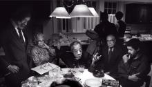 James Baldwin seated and leaning on left elbow at a table surrounded by people listening to him 