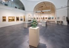 Large gallery room with white walls and gray floor tiles. Paintings hang on the walls and in the foreground is a green ceramic sculpture on a pedestal.