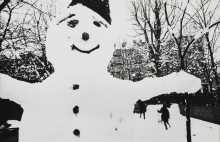 Snow man with coal button nose and eyes and sticks for eyebrows and a mouth stands in a snowy wooded park. Children run around in the background.