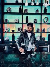 A silhouetted bar with liquor bottles and a green hue on the back wall. Behind the bar is Keith Jones II wearing a black suit jacket, white shirt, and a black brimmed hat turned backward. He is offering a wine glass full of red wine to the camera.